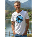 Man wearing a white t-shirt with a Tai Chi graphic design on a blue circle design standing outdoors by a lake and mountains.